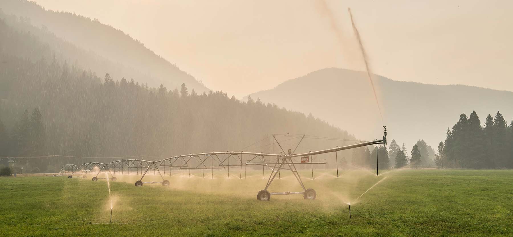 An irrigation system waters a field with barely-visible mountains in the background and the sky filled with wildfire smoke.